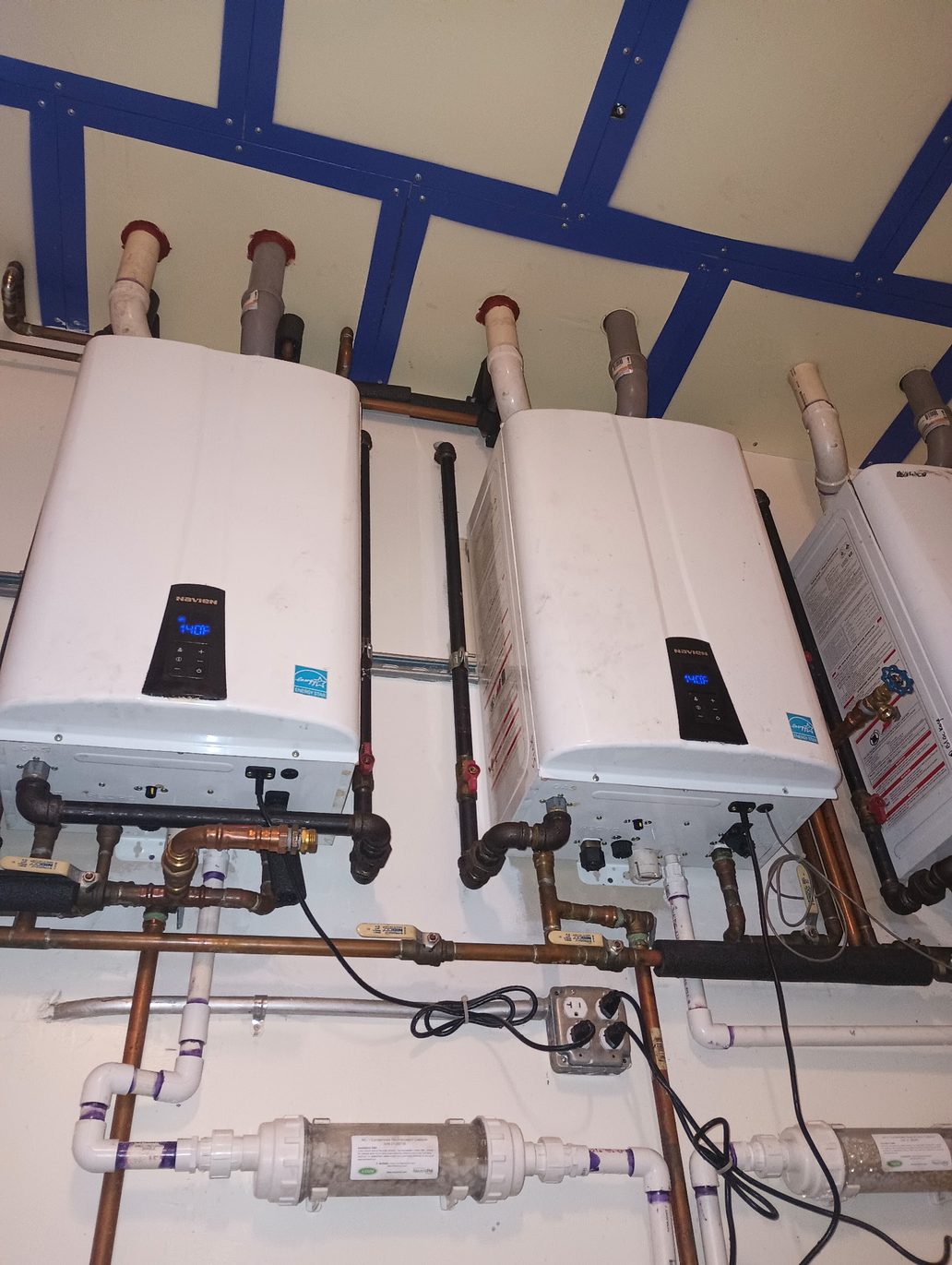 Three white Navien tankless water heaters mounted side by side on a wall, each with its own PVC intake and exhaust vent through the ceiling, copper gas and water piping with yellow-handled valves below, and two condensate neutralizer cartridges on the drain lines
