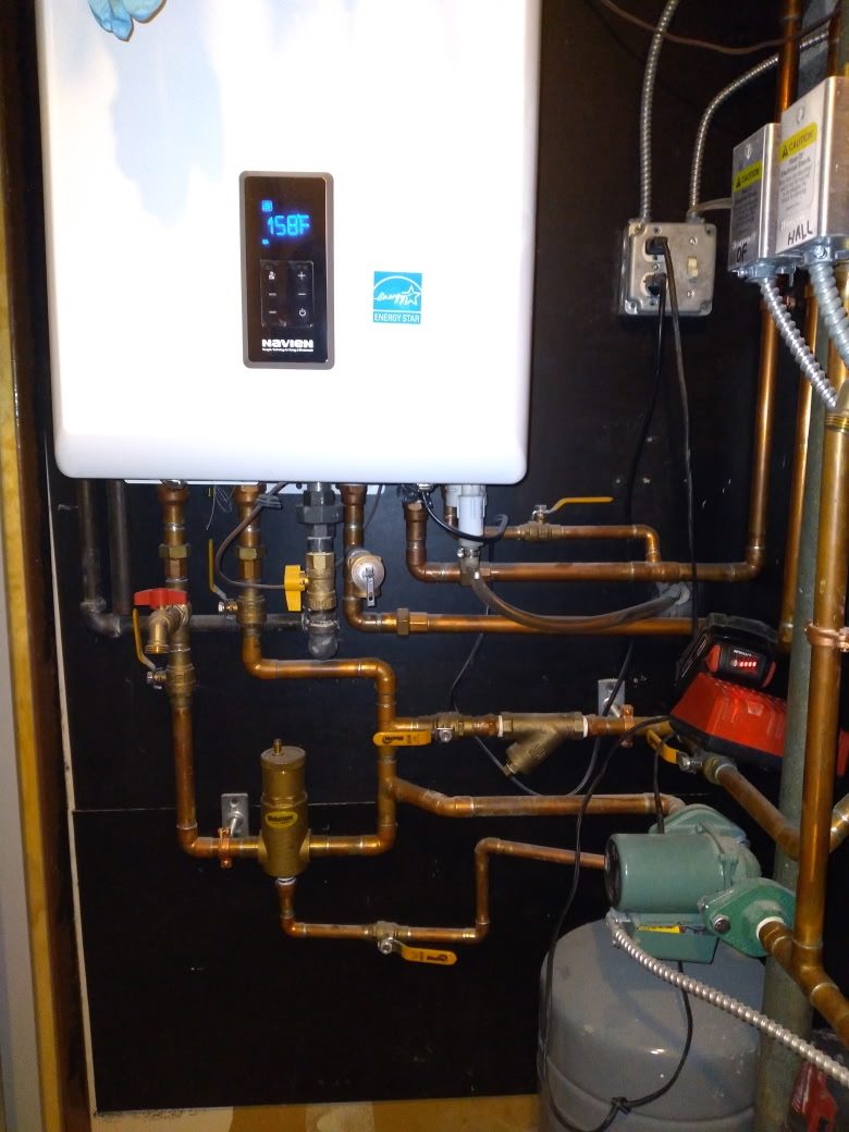 White Navien wall-mounted condensing boiler with its control panel reading 158°F, new copper piping with yellow-handled shutoff valves, a brass air separator, a circulator pump and a grey expansion tank below